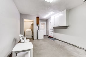 A narrow hallway with white walls and a concrete floor. There is a white chair on the left, a white door at the end of the hallway, and a white cabinet on the right.
