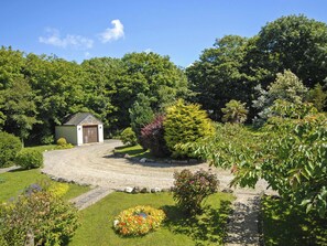 Un jardin avec un chemin de gravier, un petit abri et divers arbres et arbustes.