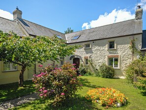 Une maison en pierre avec un toit en pente, un petit jardin fleuri et un arbre.