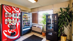 Un espace où sont installés des distributeurs automatiques de Coca-Cola et de Sprite, ainsi qu’une plante en pot et une fenêtre avec des stores.