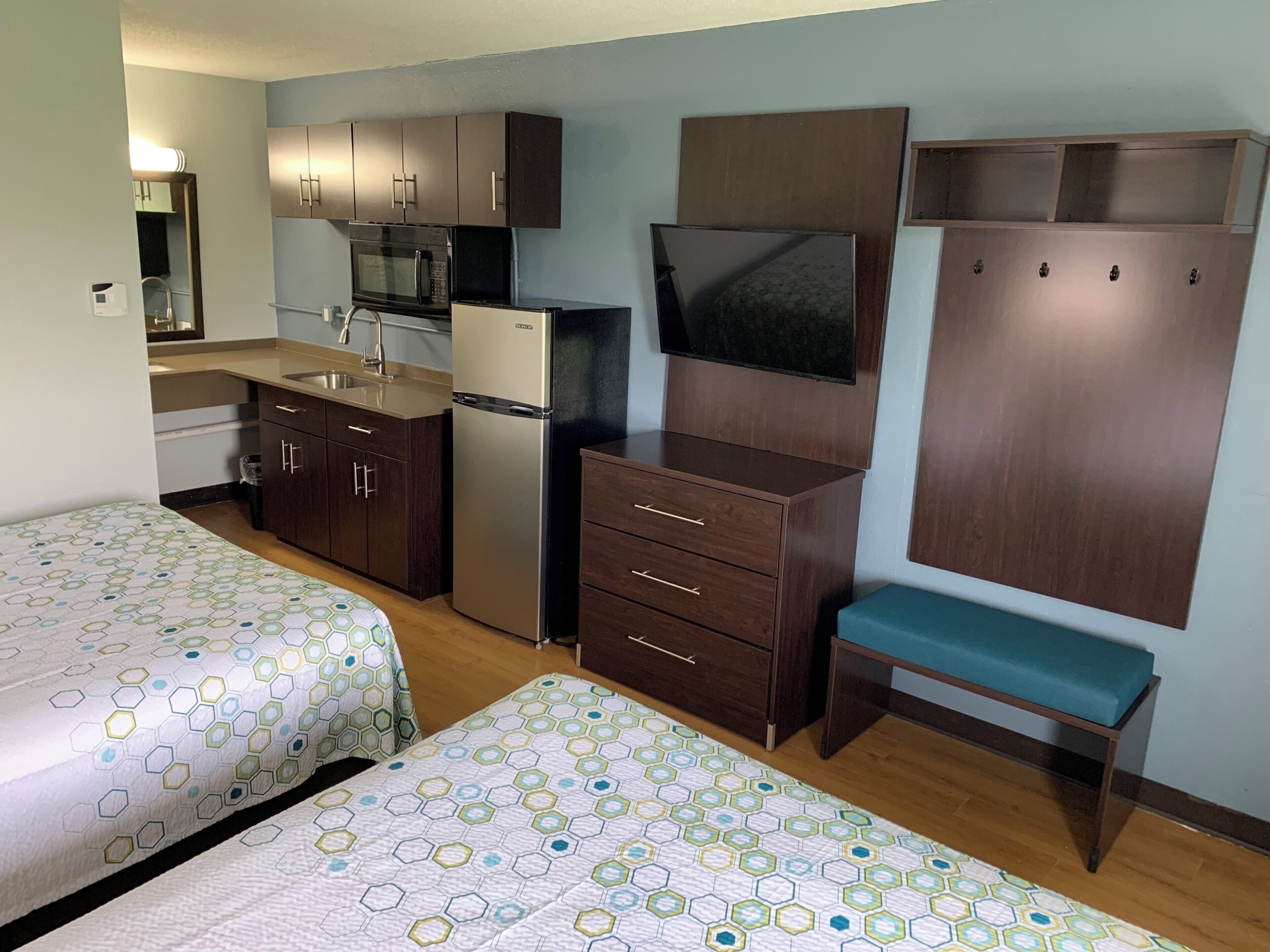 A compact kitchenette with a microwave, refrigerator, and a flat-screen TV. There are two beds with patterned bedding and a bench with a cushion.
