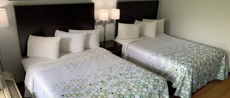 A hotel room with two beds, each with a white bedspread and matching pillows. There are two bedside tables with lamps, and a large abstract painting above the beds.