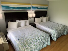 A hotel room with two beds, each with a white bedspread and matching pillows. There are two bedside tables with lamps, and a large abstract painting above the beds.