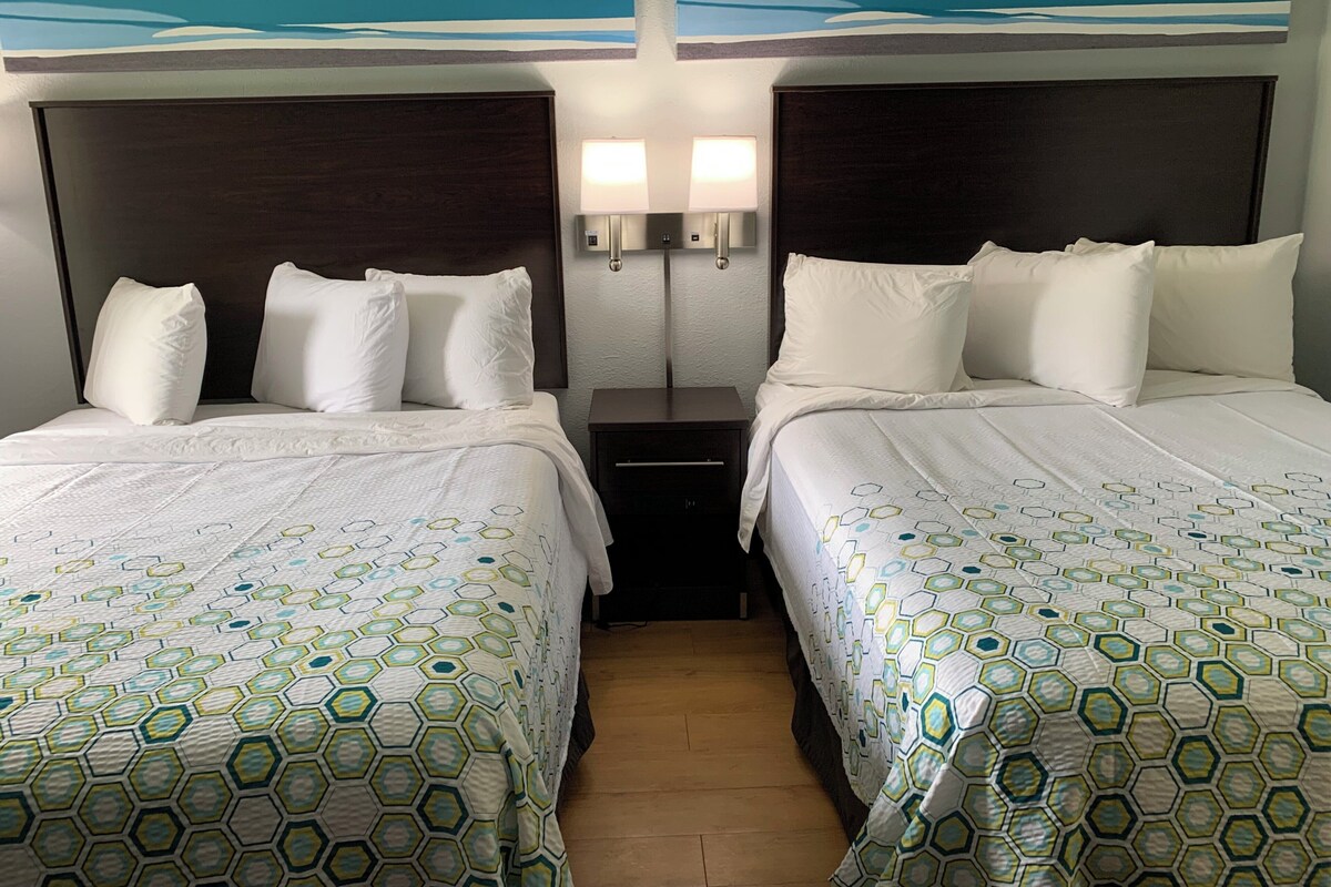 A hotel room with two beds, each with white bedding and a geometric-patterned duvet cover. There are two bedside tables with lamps, and a wall-mounted light fixture above the beds.