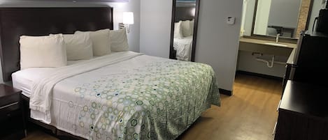Habitación de hotel con cama, mesita de noche, lámpara, espejo y baño con lavabo y espejo.