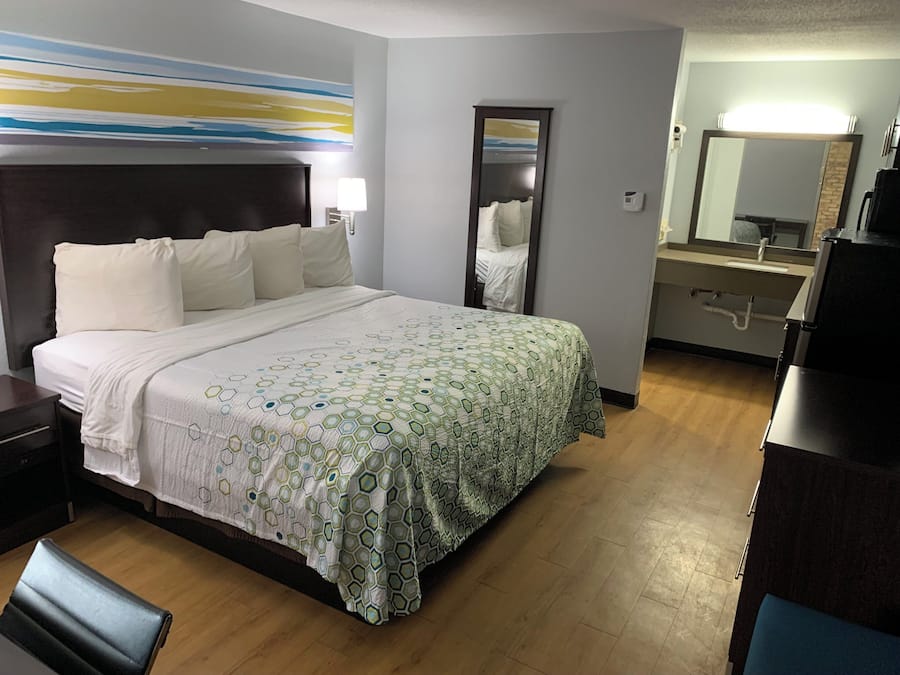 Habitación de hotel con cama, mesita de noche, lámpara, espejo y baño con lavabo y espejo.