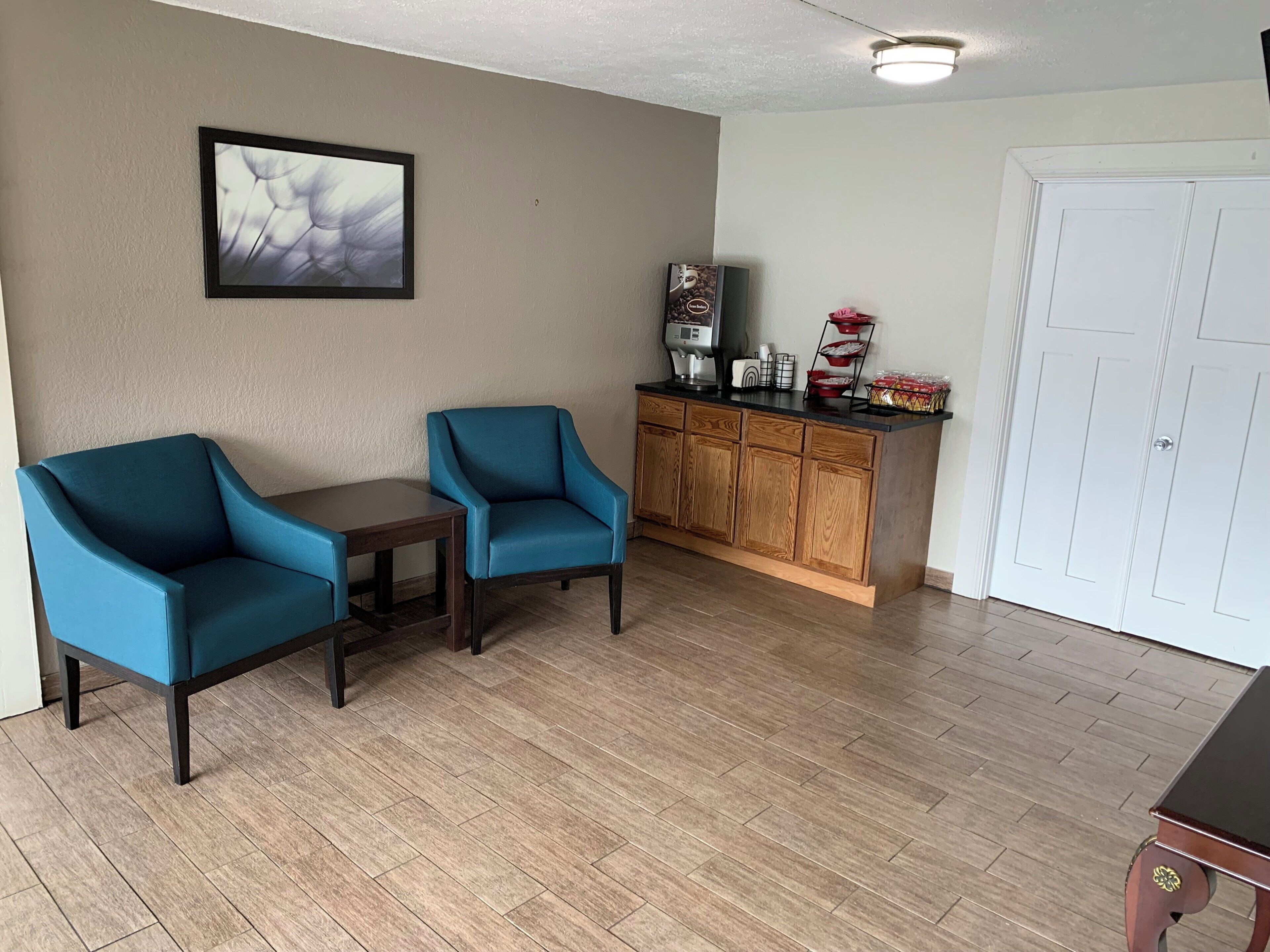 A room with two blue armchairs, a small table, a wooden cabinet with kitchen appliances, and a white door.