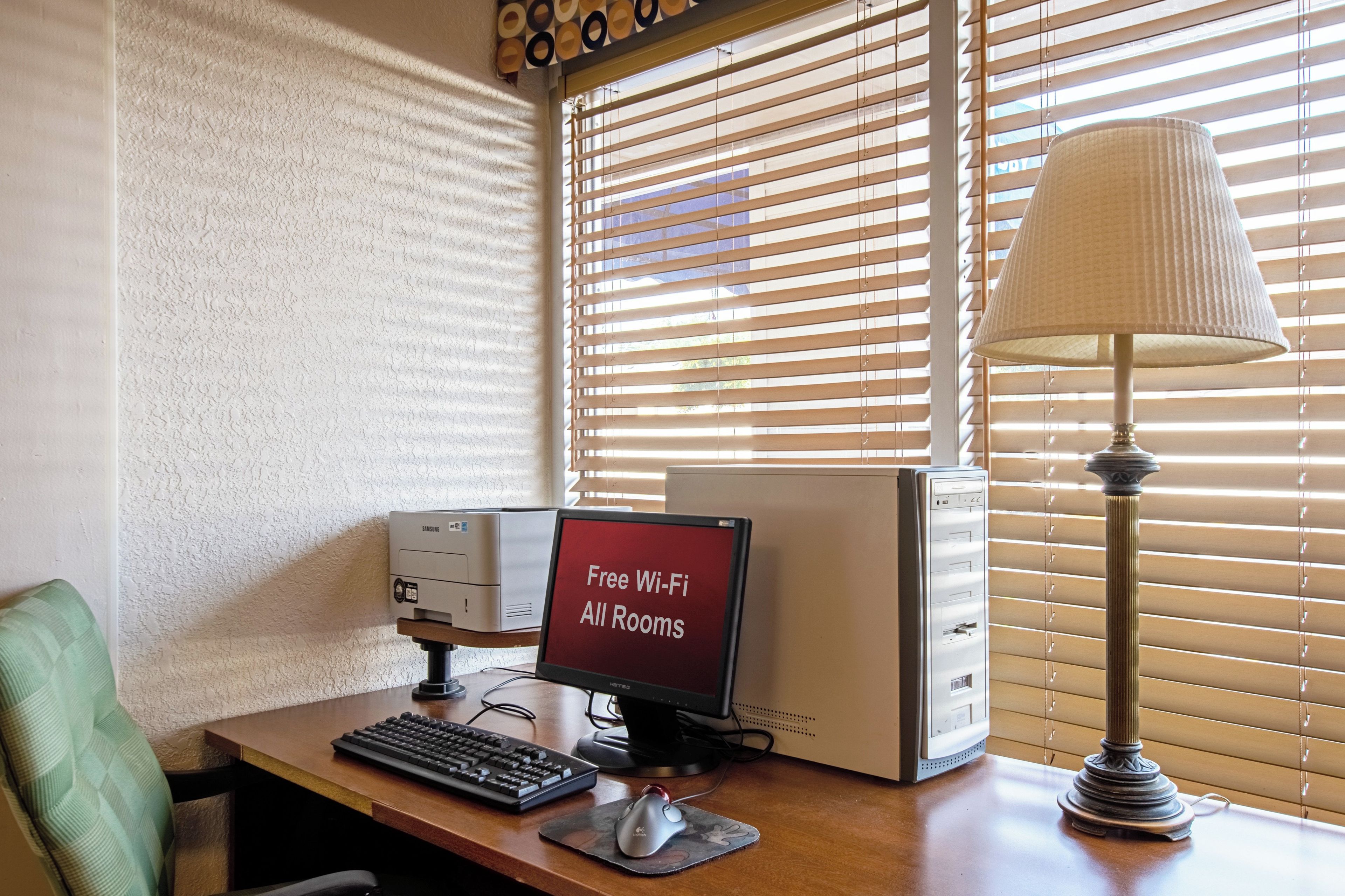A computer desk with a monitor displaying "Free Wi-Fi All Rooms," a keyboard, a mouse, and a lamp.