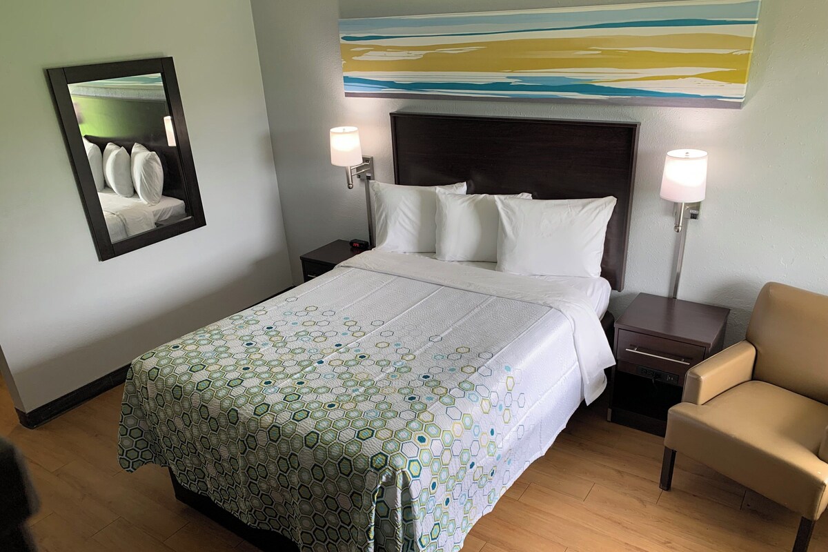 A hotel room with a bed, two bedside lamps, a chair, a nightstand, a mirror, and a painting on the wall.