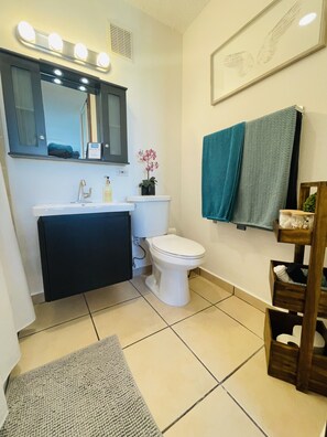 A bathroom with a white sink, a white toilet, a mirror with lights, a framed picture on the wall, and a towel rack with a towel. - Cozy and Peaceful place-Rio Grande-El Yunque Rainforest  (Río Grande)