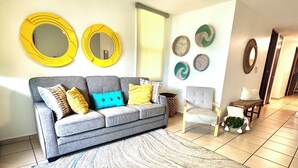 Ein Wohnzimmer mit grauem Sofa, gelb-blauen Kissen und einem grauen Sessel. An der Wand befinden sich zwei runde, gelbe Spiegel und ein kleiner Holztisch mit einer Pflanze.