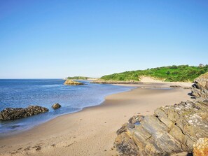 A coastal beach with clear skies, rocky formations, and a distant house on a hill. - Bwthyn yr Ardd-QC1198 (Criccieth)