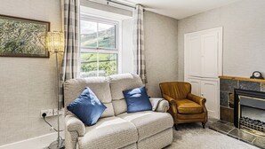 Ein gemütliches Wohnzimmer mit Sofa, Sessel und Kamin. Ein Fenster bietet Blick ins Freie.
