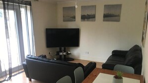 A living room with a black sofa, a wooden dining table with a potted plant, and a flat-screen TV mounted on the wall.