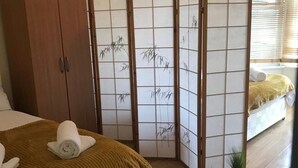 A bedroom with a bed, wooden wardrobe, shoji screens, a small plant, and a window with blinds.
