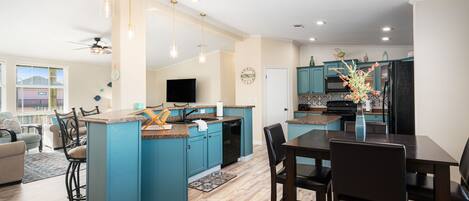 Een moderne keuken met blauwgroene kastjes, een centraal eiland en een eethoek met donker houten meubels.