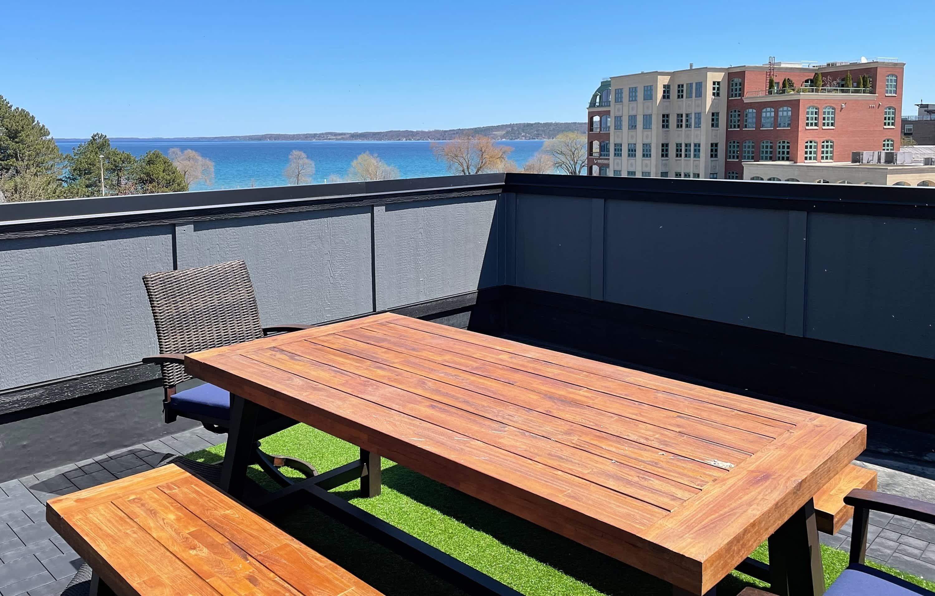 Ein Holztisch und eine Bank auf einer Terrasses mit Blick auf das Meer und Gebäude im Hintergrund.