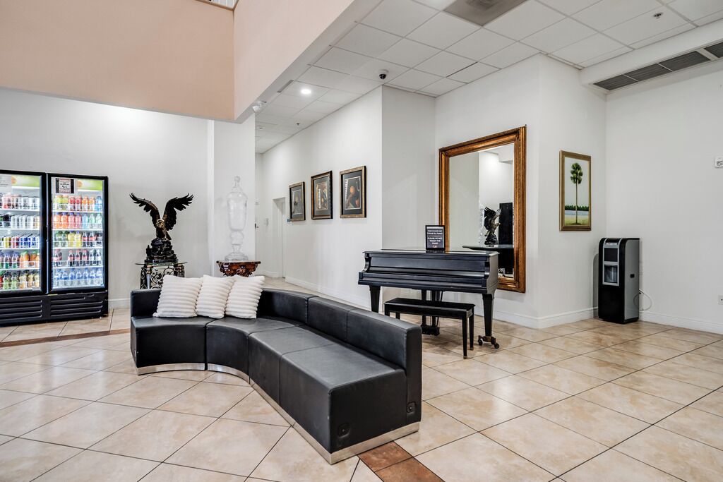 Un salon moderne avec un canapé en cuir noir aux lignes courbes, un grand piano et un réfrigérateur rempli de boissons.