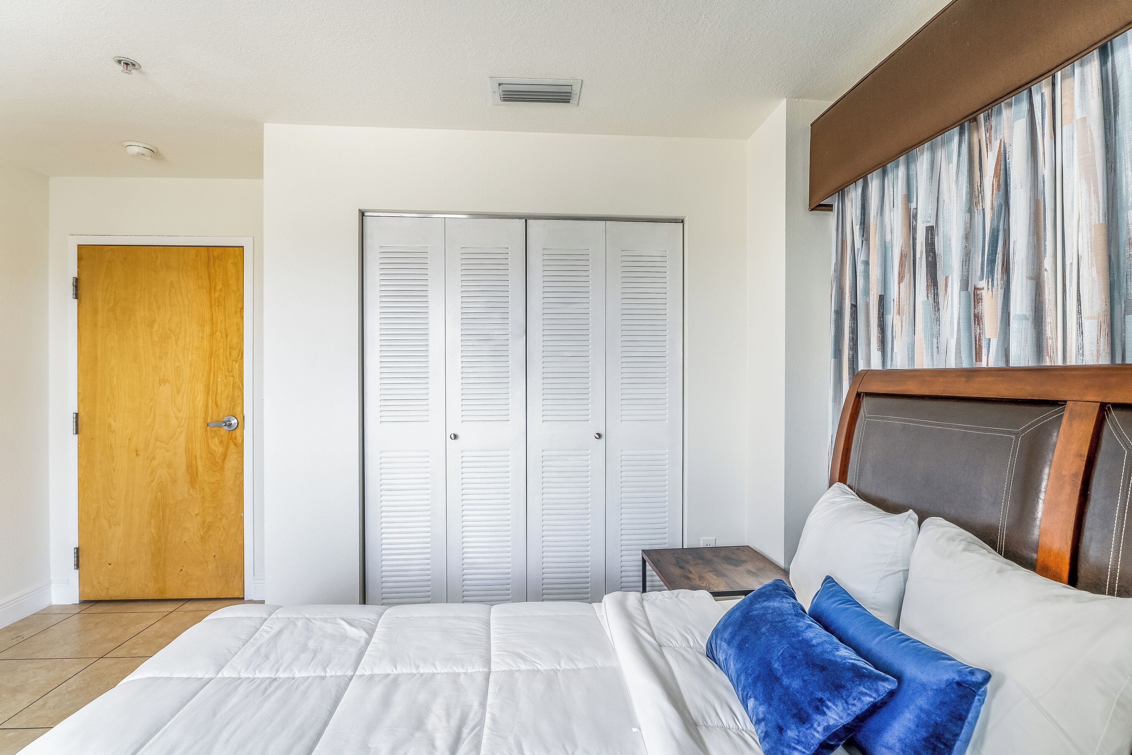 A bedroom with a bed, a wooden door, a closet with white louvered doors, and a headboard with a wooden frame.
