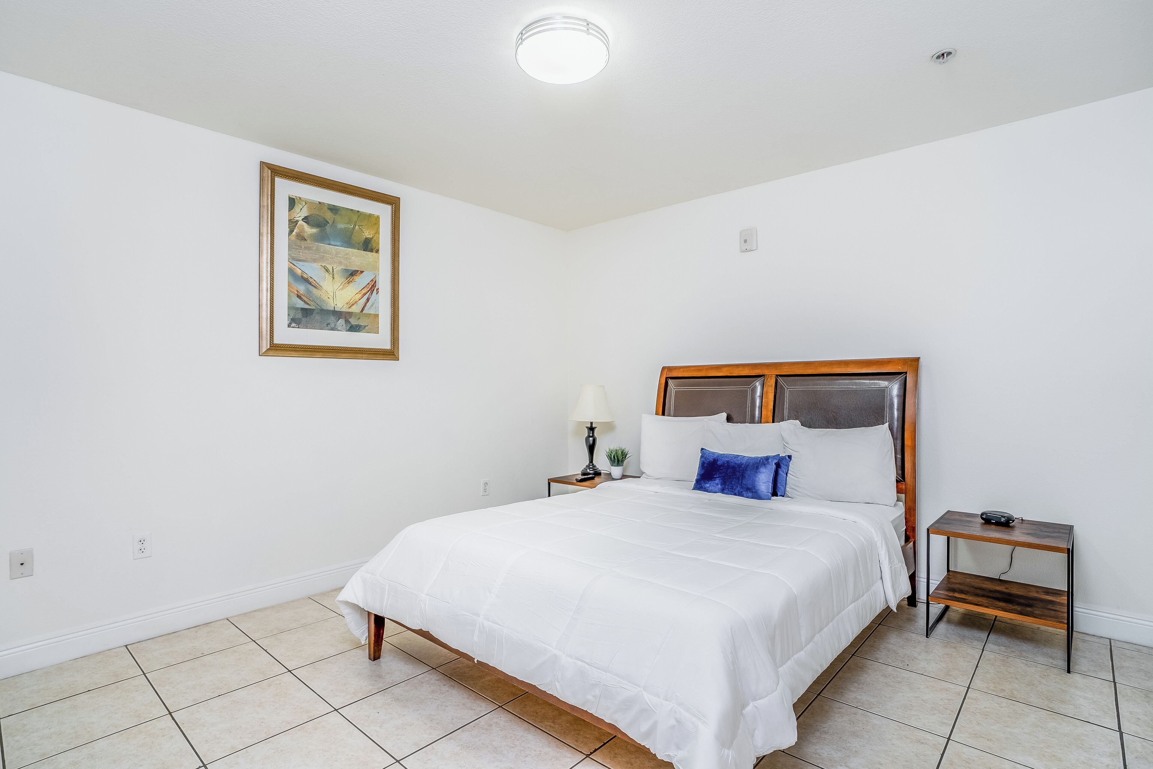 Een keurig opgemaakte slaapkamer met een groot bed, een nachtkastje met een lamp en een lijst met een foto aan de muur.