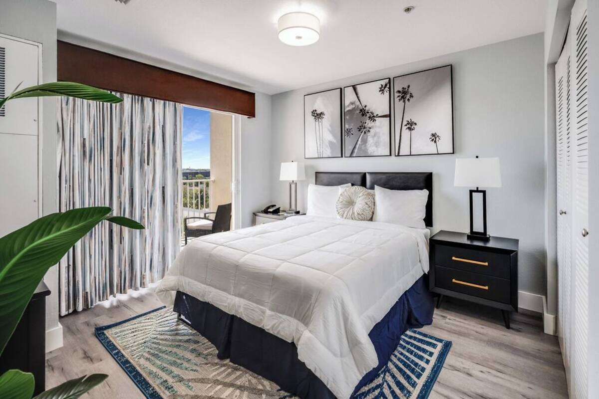A modern bedroom with a large bed, a nightstand, a desk, and a view of the outdoors.
