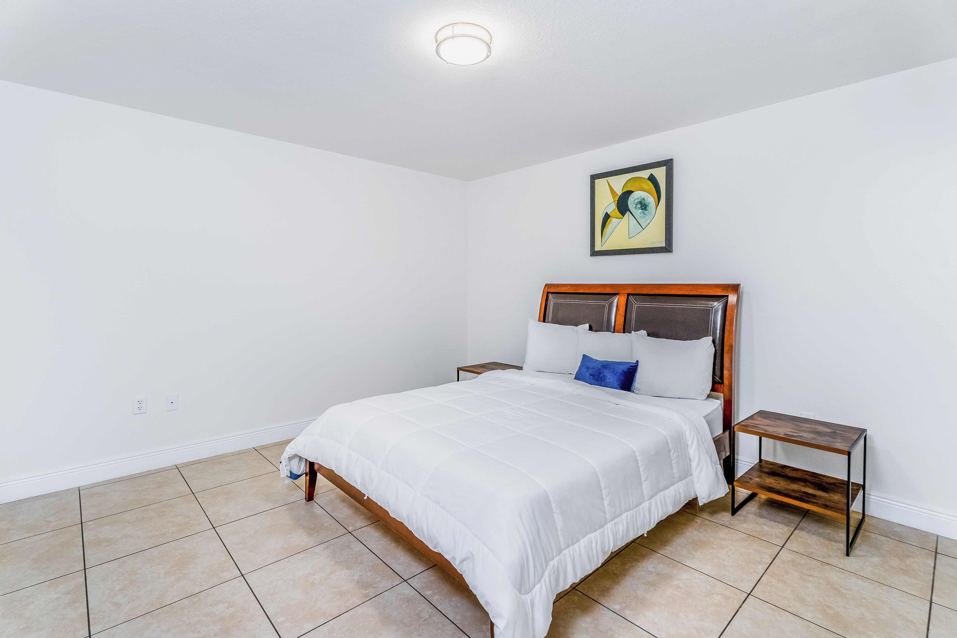 A bedroom with a bed, nightstands, a painting, and tiled flooring.