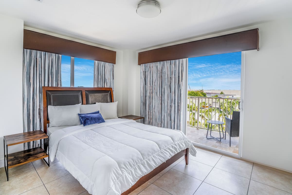 A hotel room with a large bed, two nightstands, a view of the outdoors, and a balcony with a table and chairs.