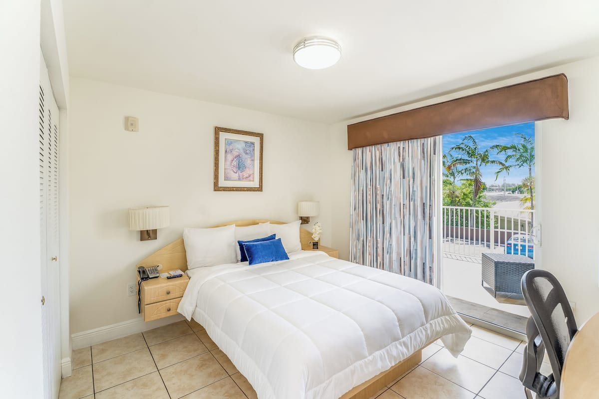 A hotel room with a bed, bedside tables, a lamp, a painting, and a view of palm trees through the window.
