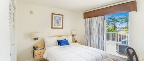 Una camera d'albergo con un letto, comodini, una lampada, un quadro e vista su palme attraverso la finestra.