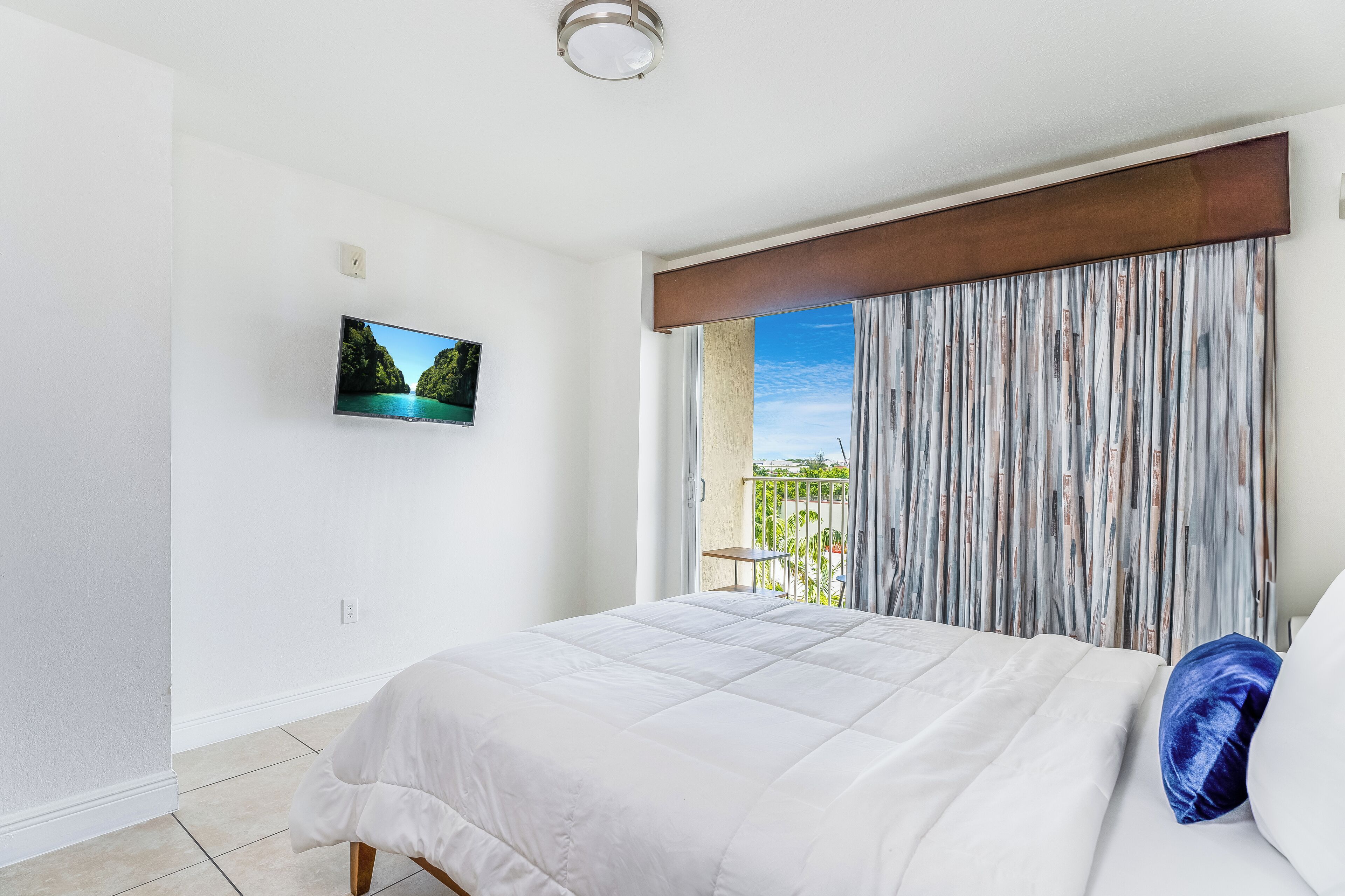A hotel room with a bed, a television on the wall, and a view of the outdoors through a window.