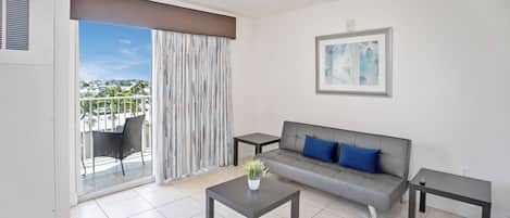 A modern living room with a grey sofa, a small table with a plant, and a view of a balcony with a chair.