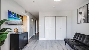 A modern living room with a black leather sofa, a flat-screen TV on a black cabinet, a large potted plant, and a white closet with sliding doors. - South River Suites (Miami)
