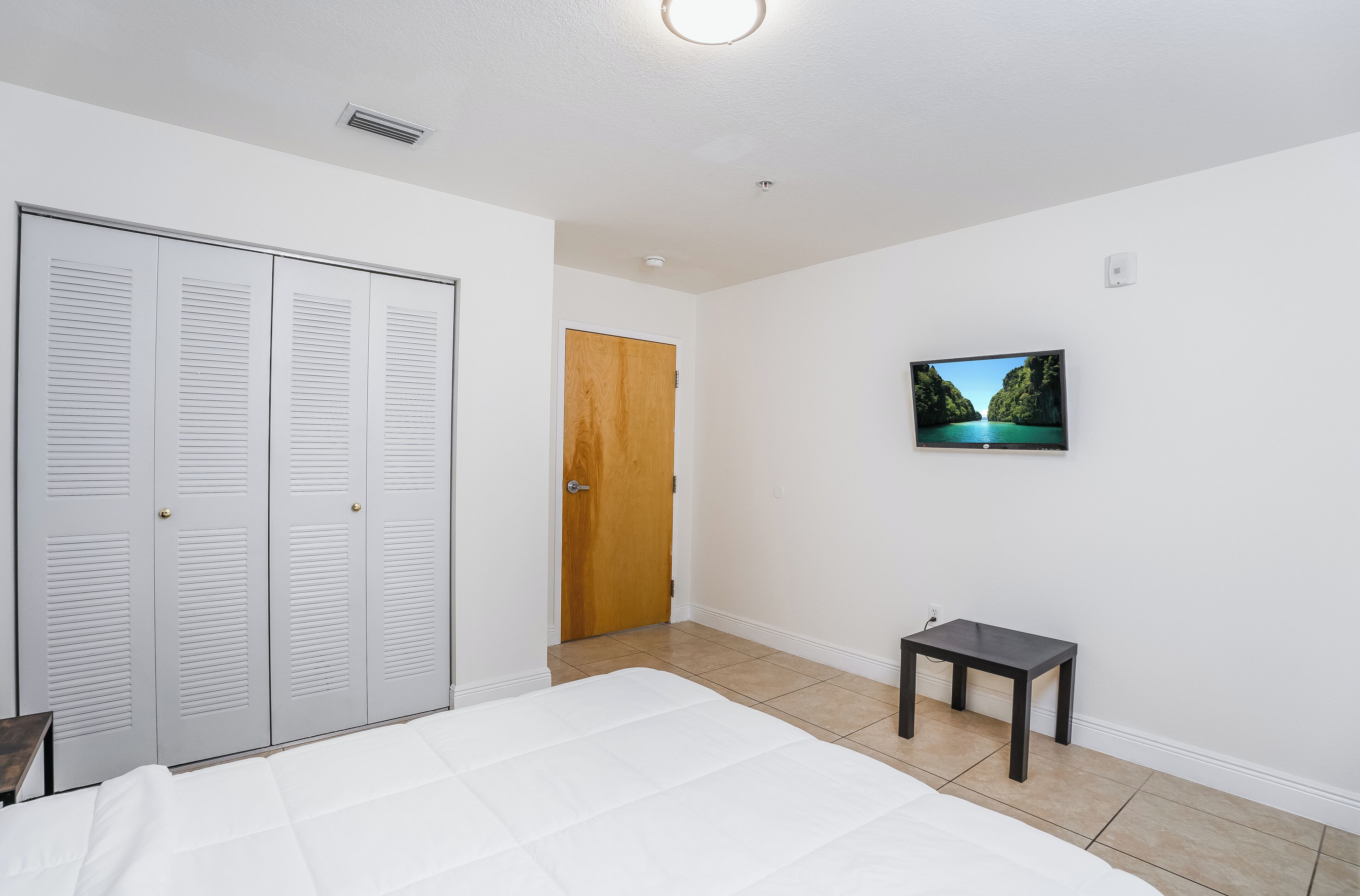 Un dormitorio con una cama, una puerta de madera, una mesita pequeña y un televisor fijado a la pared.