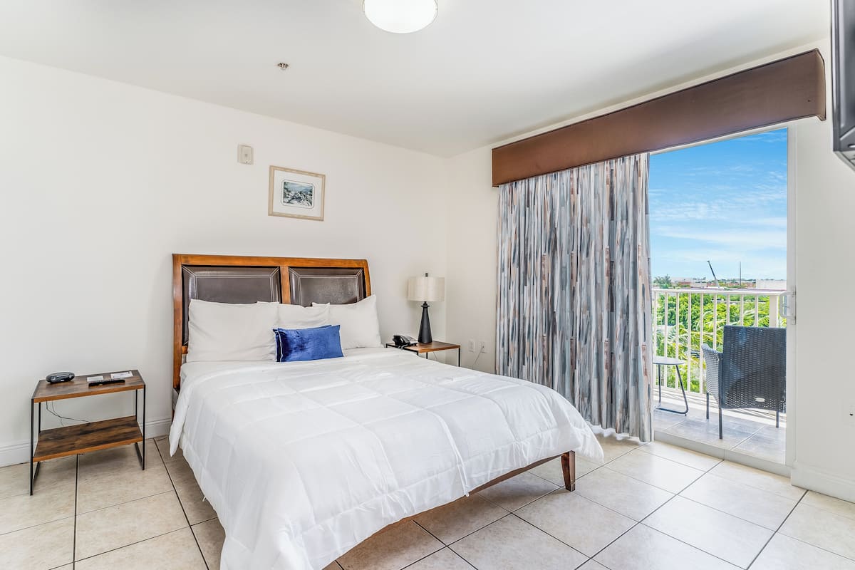 A hotel room with a large bed, a bedside table with a lamp, a small table with a phone, and a view of a balcony with outdoor furniture.