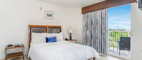 A hotel room with a large bed, a bedside table with a lamp, a small table with a phone, and a view of a balcony with outdoor furniture.