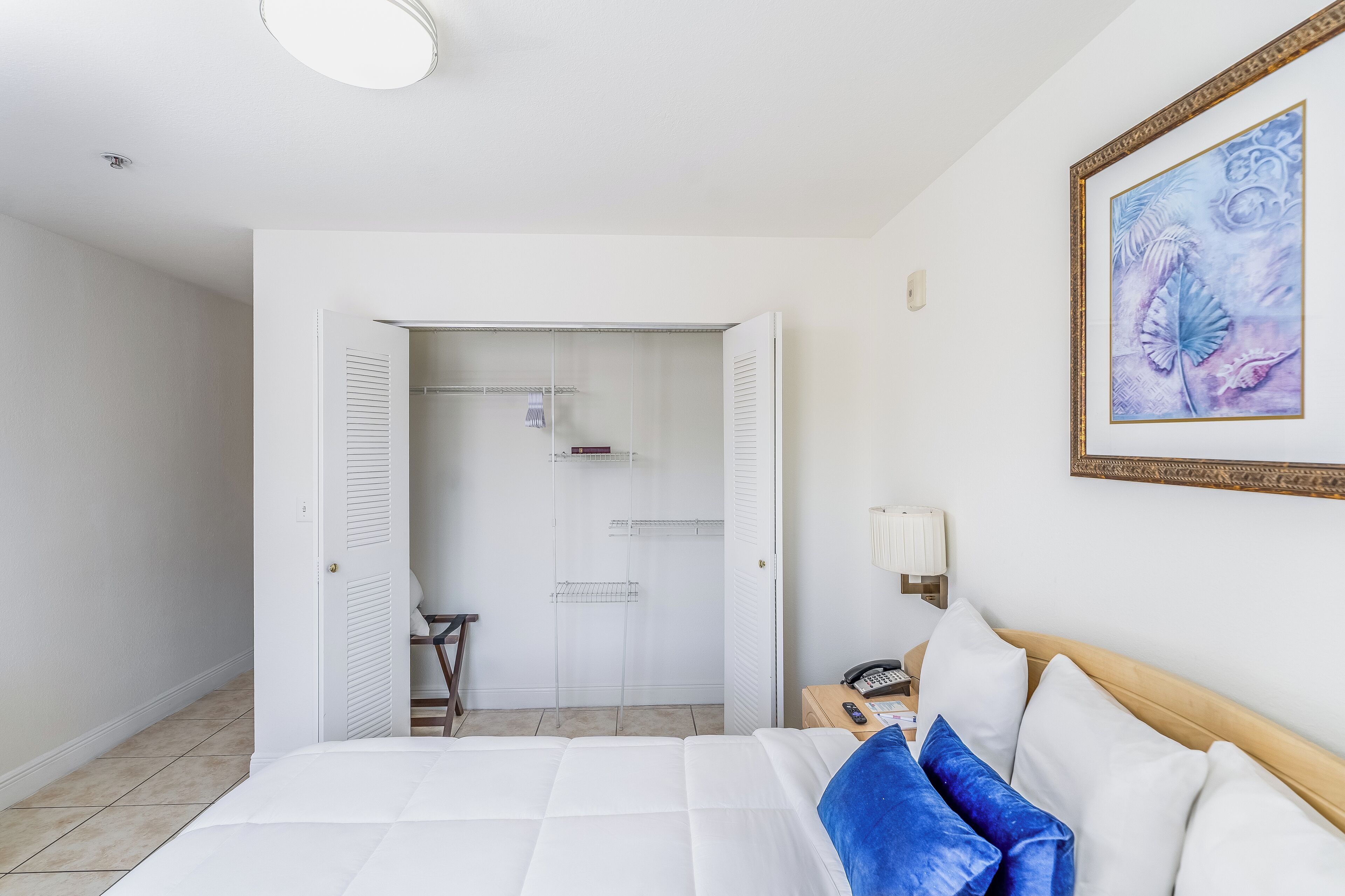 A hotel room with a bed, a nightstand, a lamp, a framed artwork, and a closet with shelves.