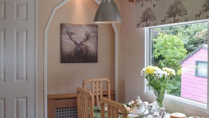 A dining area with a table set for tea, a painting of a deer, and a window with a view of greenery and a pink building. - A perfect countryside escape for lovers of the outdoors. (Ayr)
