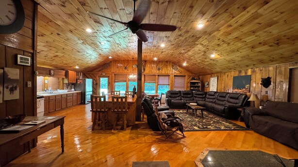 A spacious living area with a wooden ceiling, a large black leather sofa, a wooden dining table, and a kitchen visible through an open door.