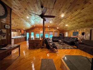 A spacious living area with a wooden ceiling, a large black leather sofa, a wooden dining table, and a kitchen visible through an open door. - Adirondack Mountain Lodge (Glenfield)