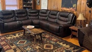 A cozy living room with a large L-shaped leather sofa, a patterned rug, wooden walls, and a ceiling with exposed beams.