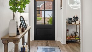 Un couloir avec un plancher en bois, une porte noire, un tapis bleu, une plante, un vase, un tableau encadré, une étagère contenant des ustensiles de cuisine et un panneau lumineux au néon.
