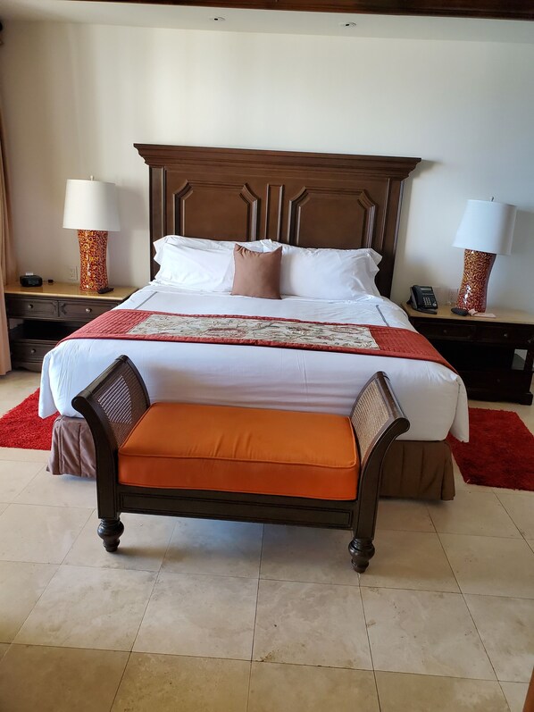 Une chambre d’hôtel avec un grand lit, une tête de lit en bois, deux tables de chevet avec des lampes, un tapis rouge et un banc au pied du lit.