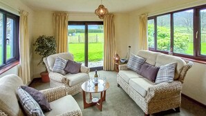 Un salón acogedor con muebles de mimbre, una mesita de centro y ventanas amplias que ofrecen vistas a un paisaje verde.