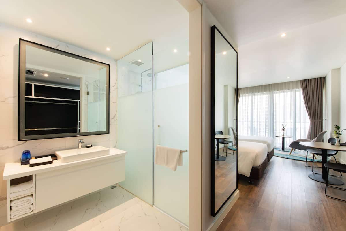 A modern hotel room with a large mirror, a white vanity with a sink, a glass shower enclosure, a bed with a white duvet, a small table with a chair, and a window with curtains.