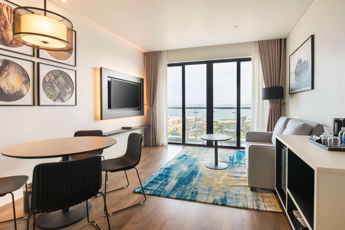 A modern hotel room with a large window, a flat-screen TV, a dining area with a round table and chairs, a sofa, and a coffee station.