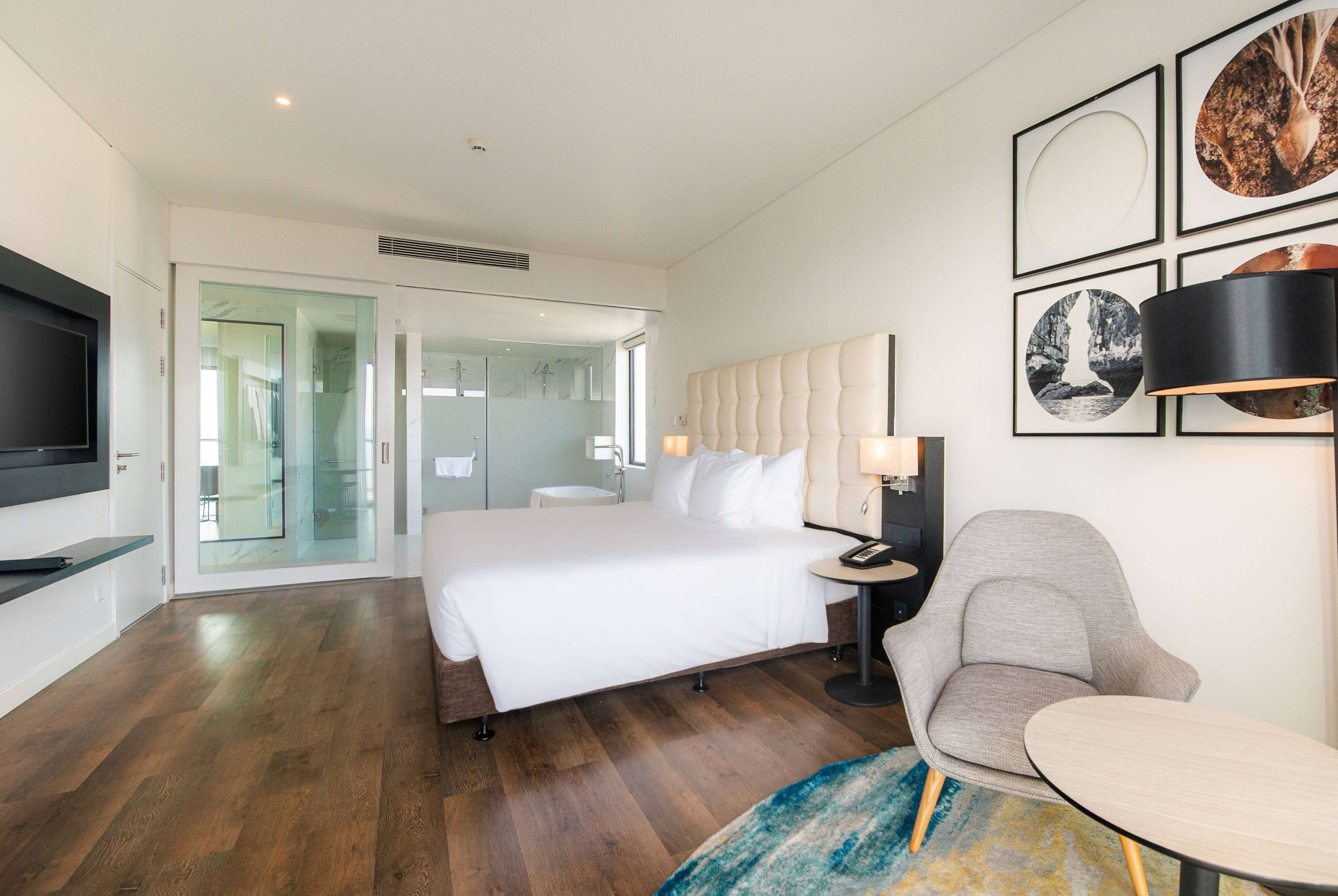 Ein modernes Hotelzimmer mit einem großen Bett, einer Sitzecke mit Sessel und Tisch, einem Fernseher und Bildern an der Wand.