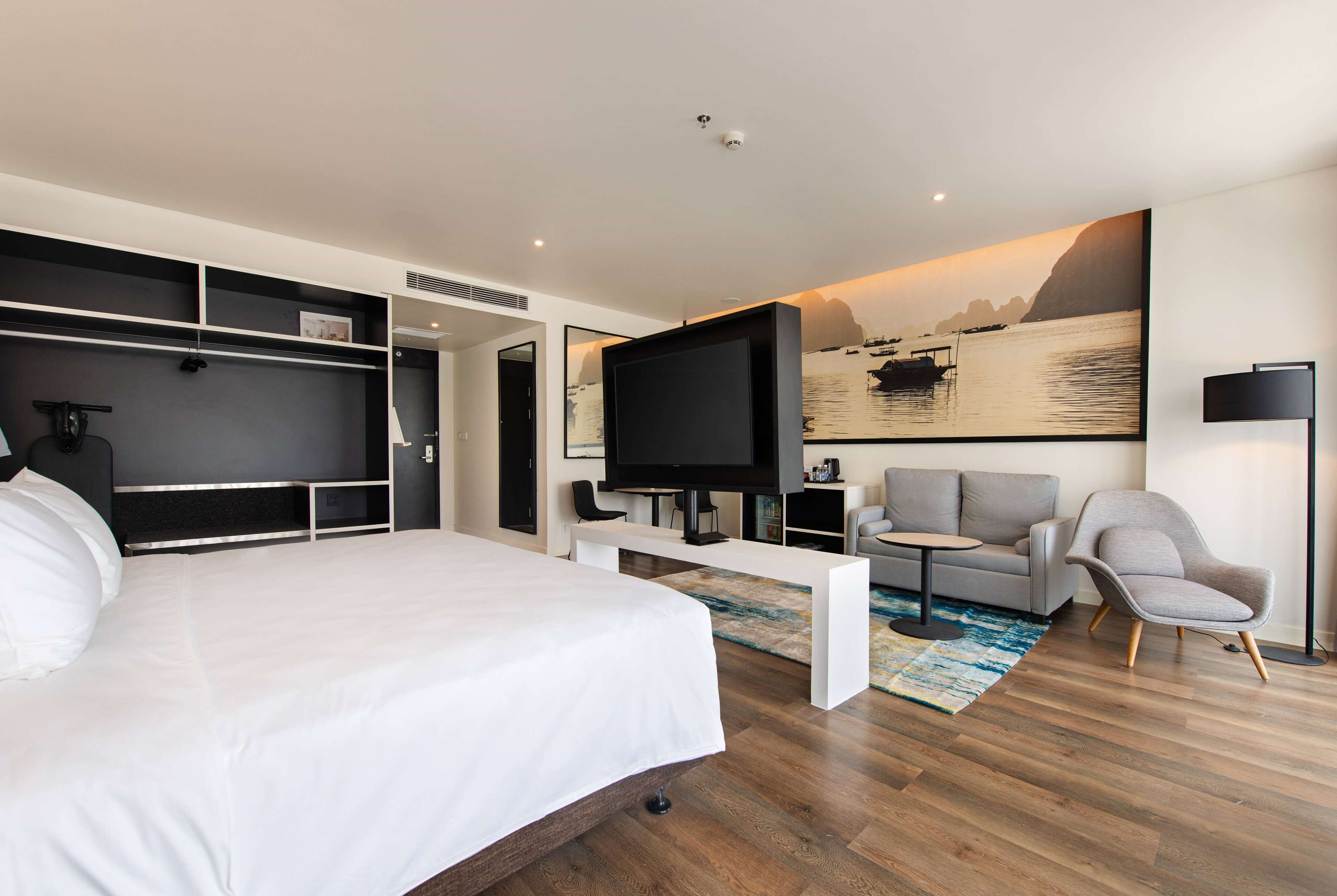 A modern hotel room with a large bed, a flat-screen TV, a sofa, and a painting of a landscape.