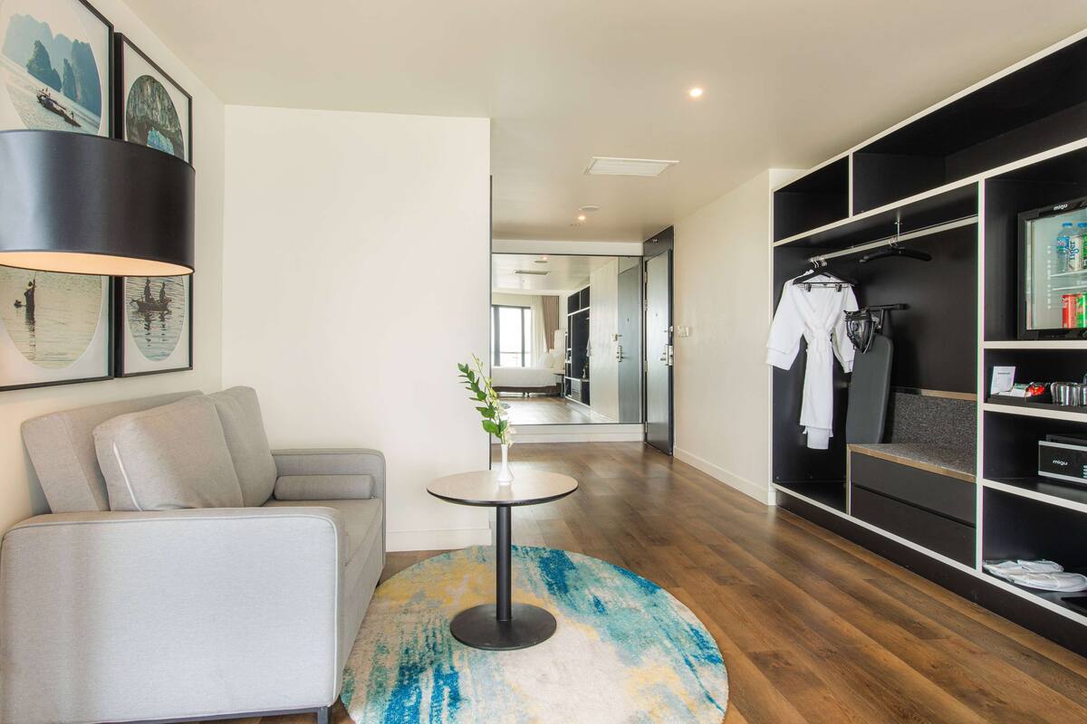A modern hotel room with a grey sofa, a round coffee table with a vase, a wardrobe with hanging clothes, and a television.