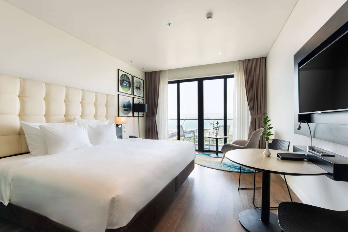 A modern hotel room with a large bed, a desk with a chair, a TV, and a balcony with a view.