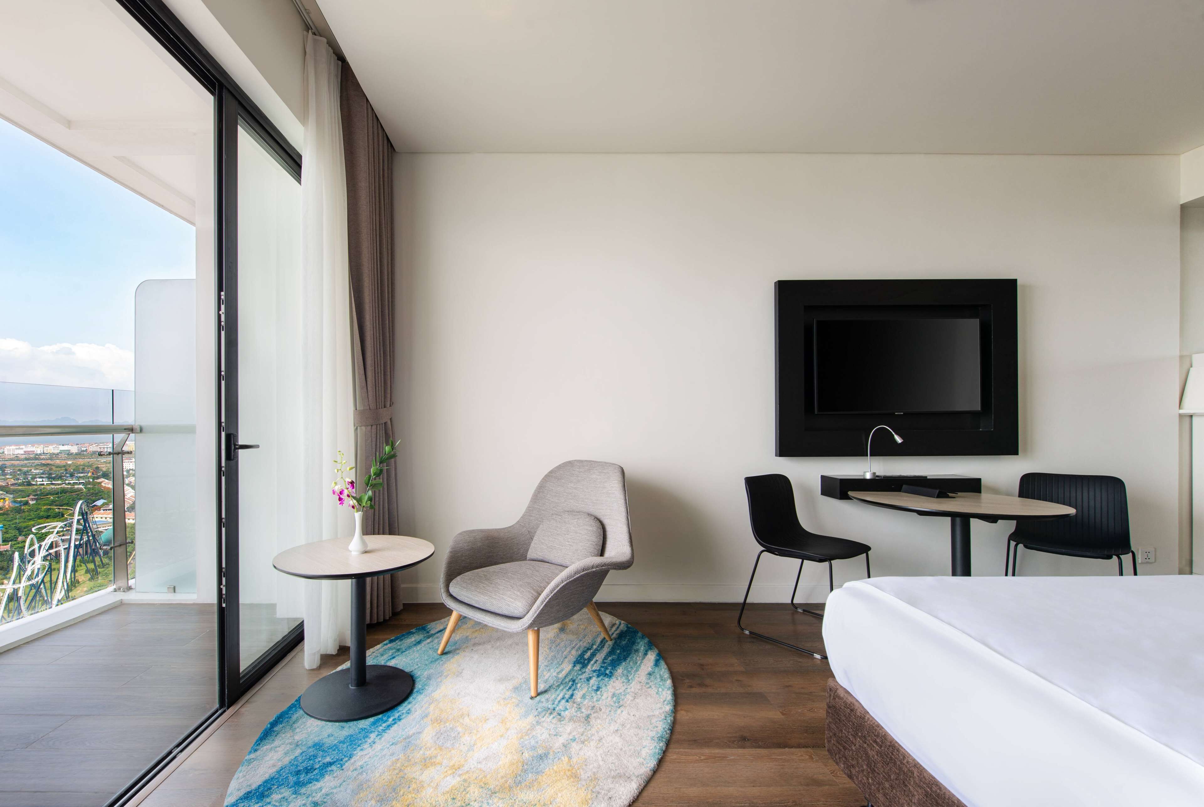 A modern hotel room with a large bed, a round table, two chairs, a TV mounted on the wall, and a balcony with a view of the city.